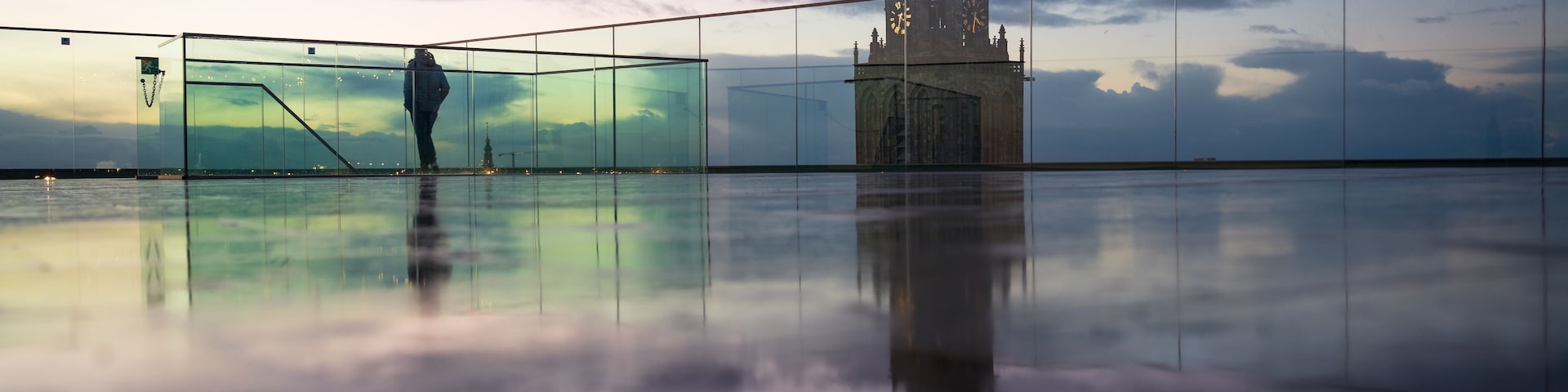 Uma pessoa caminhando sobre uma superfície refletora, com uma torre histórica ao fundo.