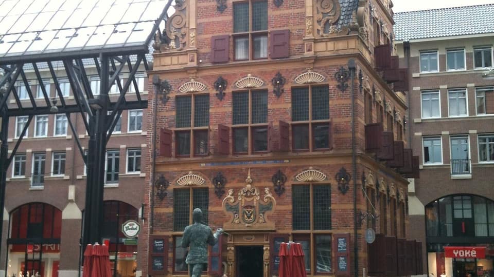 Ein historisches Gebäude mit aufwendiger Architektur, einer Statue davor und Außenbestuhlung.