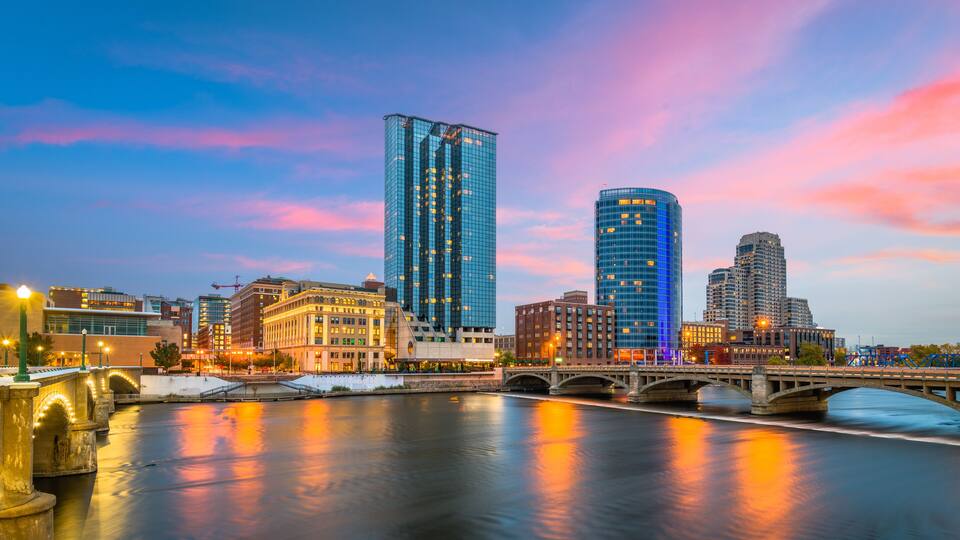 A cityscape with a river, a bridge, and modern buildings under a colorful sky.