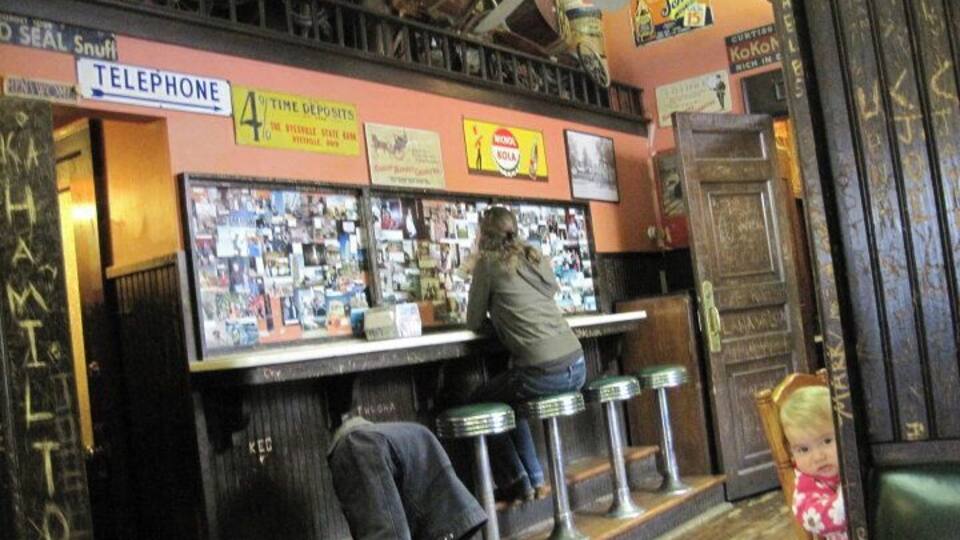 Eine Bar mit Telefon, ein Mann sitzt an der Bar und ein Kind steht in der Nähe des Eingangs.