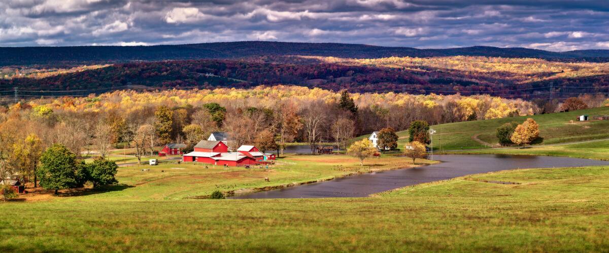 A rural landscape with a river, red barns, and autumn foliage.