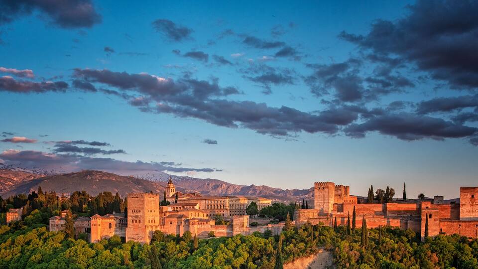 A historic city with a prominent castle, lush greenery, and mountains in the background.