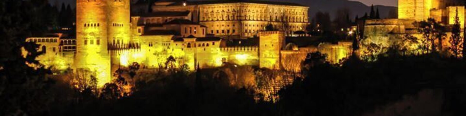 Un castillo histórico con torres y murallas iluminadas, rodeado de árboles y montañas al fondo.