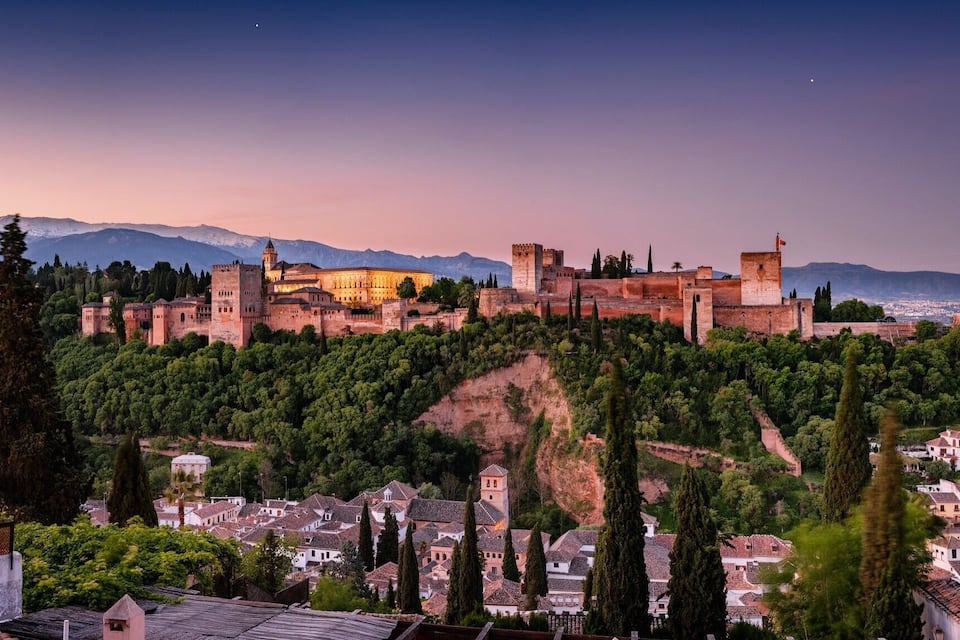A historic fortified city with a castle on a hill, surrounded by lush greenery and a backdrop of mountains.