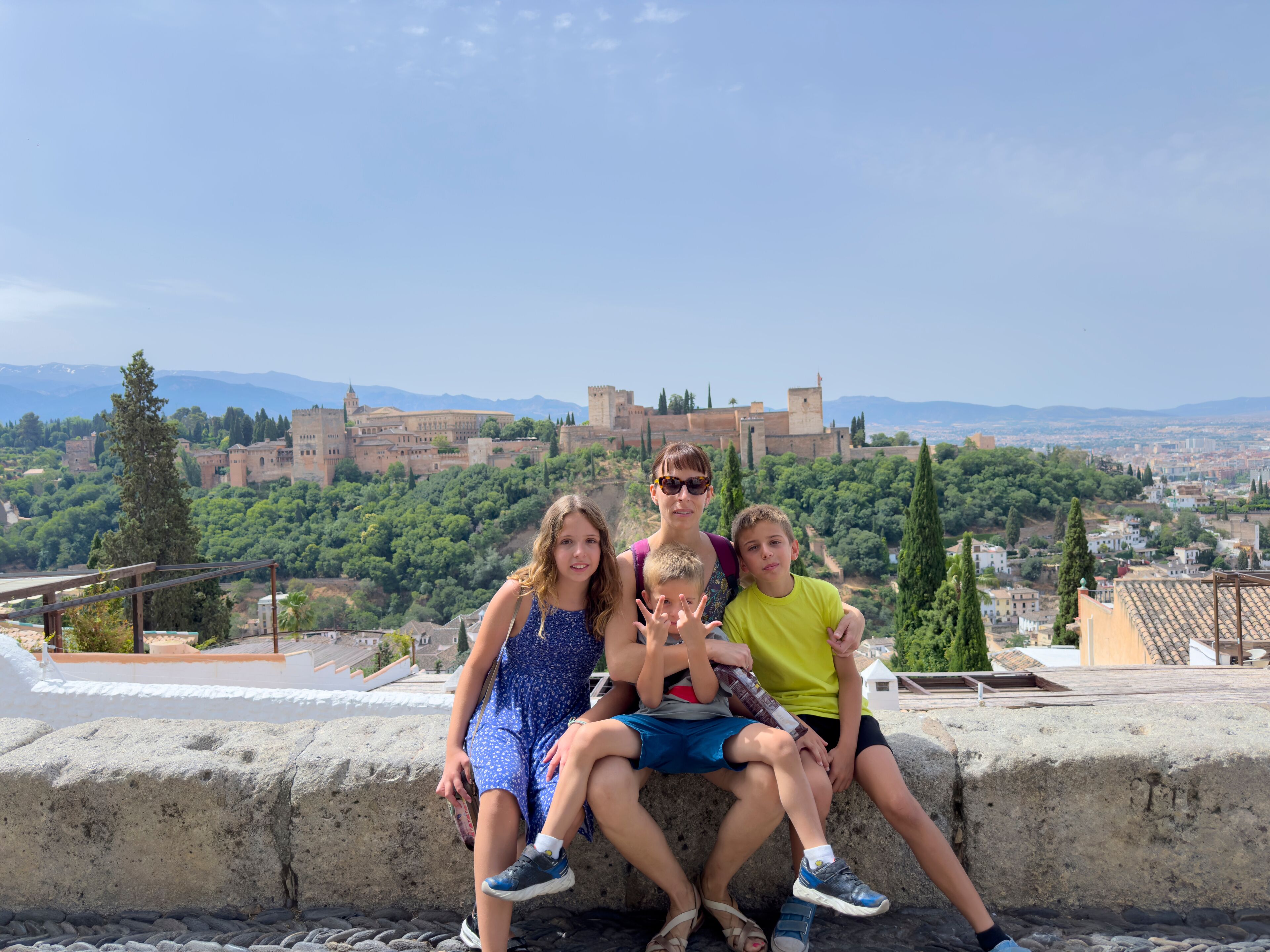 Un grupo de personas sentadas sobre un muro de piedra con vistas a una ciudad histórica, donde se distingue un castillo imponente al fondo.