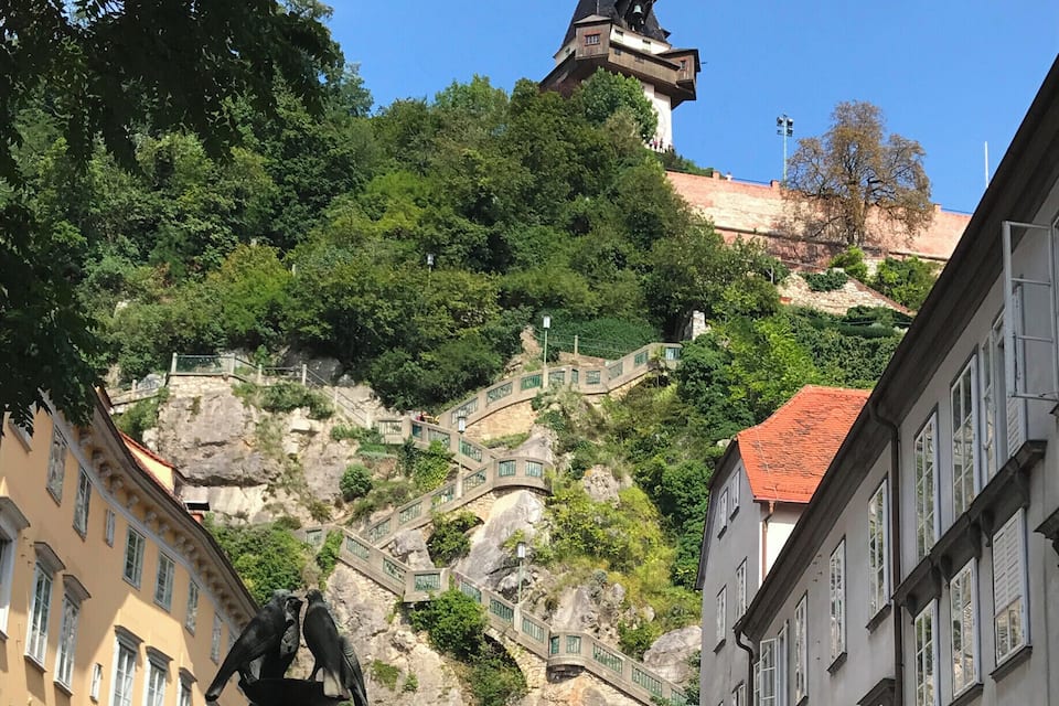 A historic town square with a fountain, a statue, and a steep staircase leading to a tower.