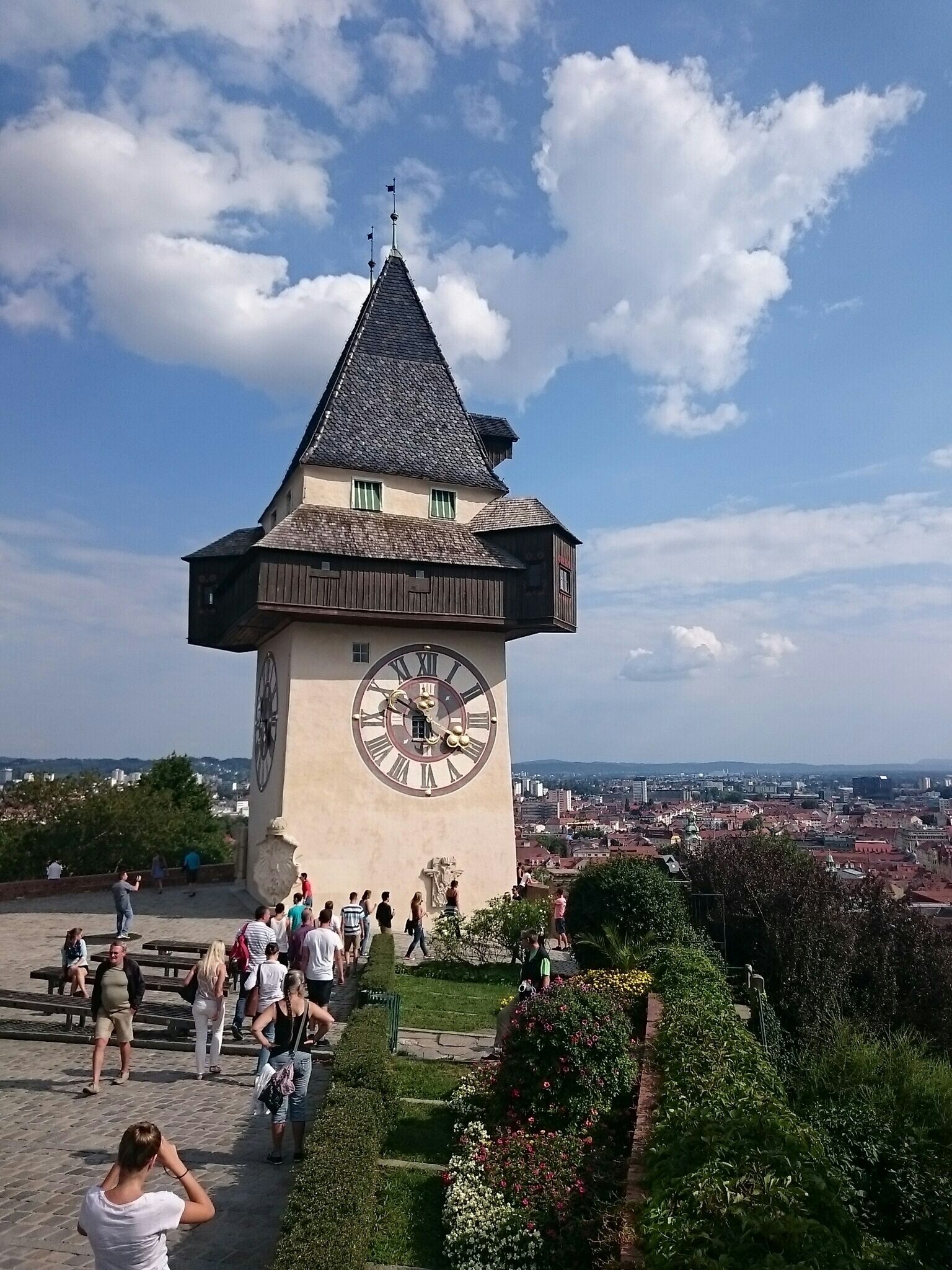 Una torre del reloj con techo a dos aguas y una esfera grande, rodeada por un jardín con flores y un paisaje urbano al fondo.