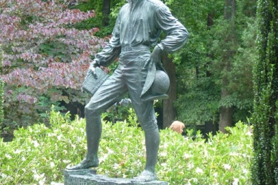 Uma estátua de um homem vestido com roupas da época, em um pedestal de pedra rodeado por vegetação.