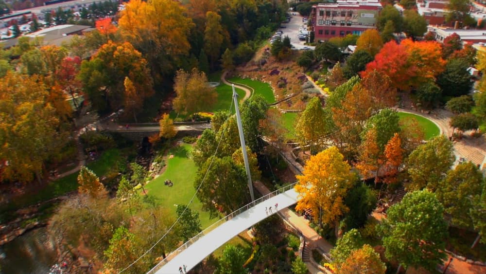 Eine Luftaufnahme eines Parks mit einer Hängebrücke, Bäumen in Herbstfarben und einem gewundenen Weg.