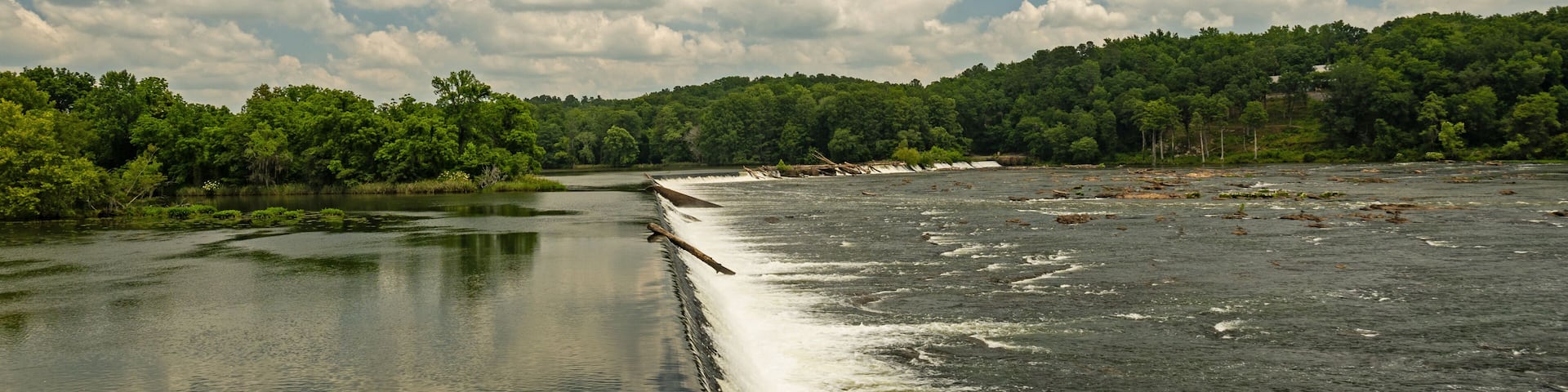A river with a dam, surrounded by dense green forest under a partly cloudy sky.
