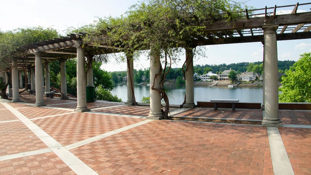 A brick-paved walkway with a pergola structure, columns, and a view of a river with houses in the background.