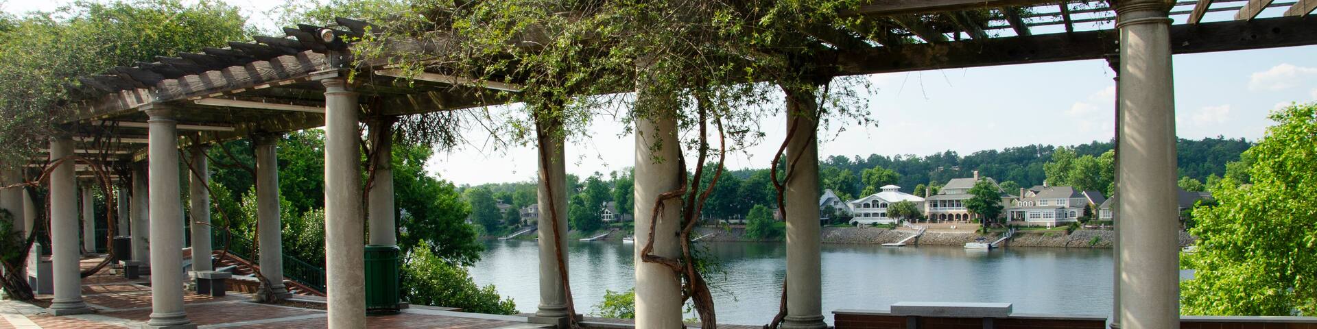 A section of a river walk with a shaded rest area under a pergola, with white columns. There is a view of the river.