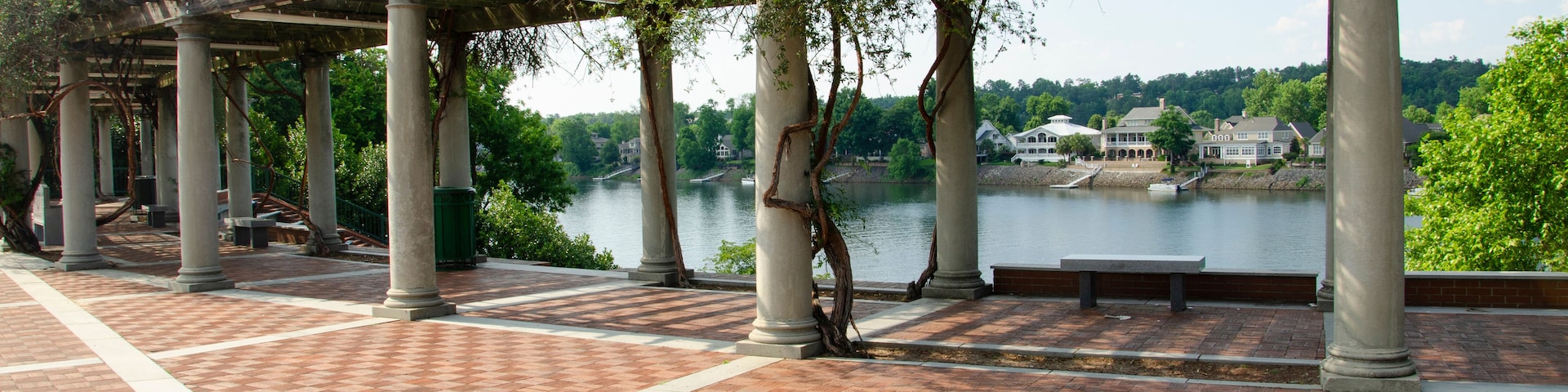Un sendero empedrado de ladrillo con una estructura de pérgola, columnas y vista a un río con casas al fondo.