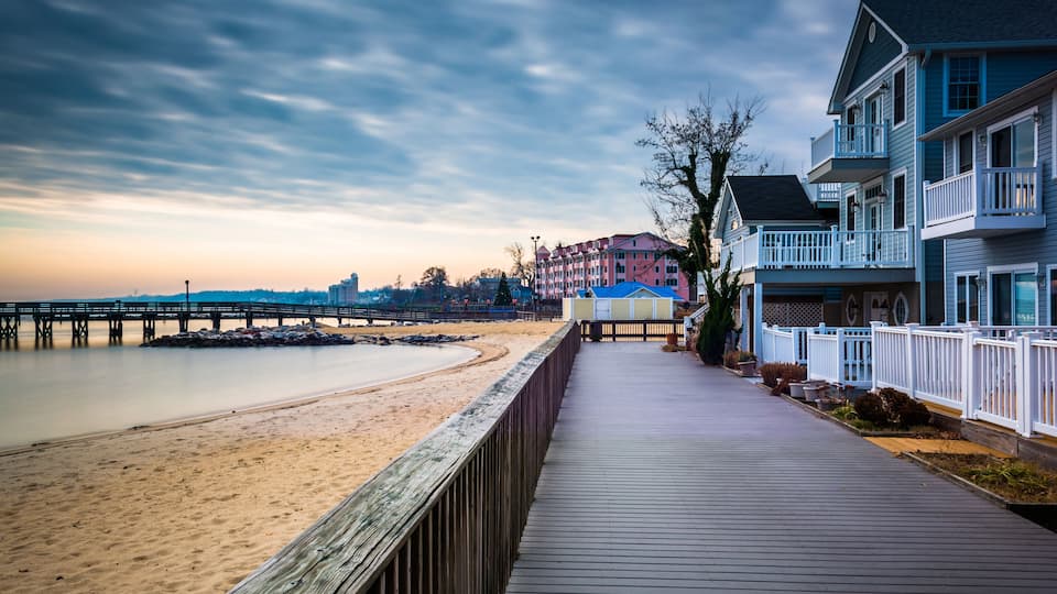 A wooden boardwalk along a sandy beach with buildings and a pier in the background.