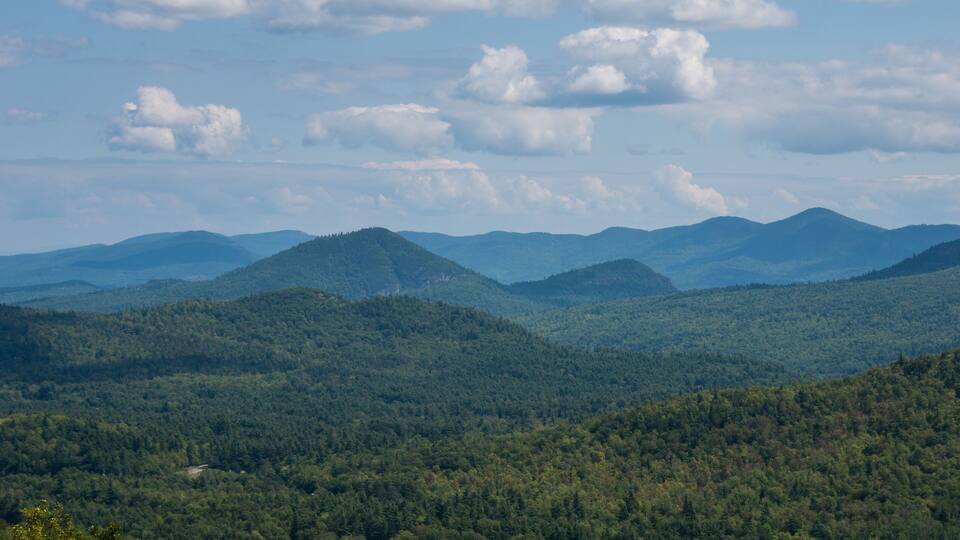 A mountainous landscape with dense forests and rolling hills under a partly cloudy sky.
