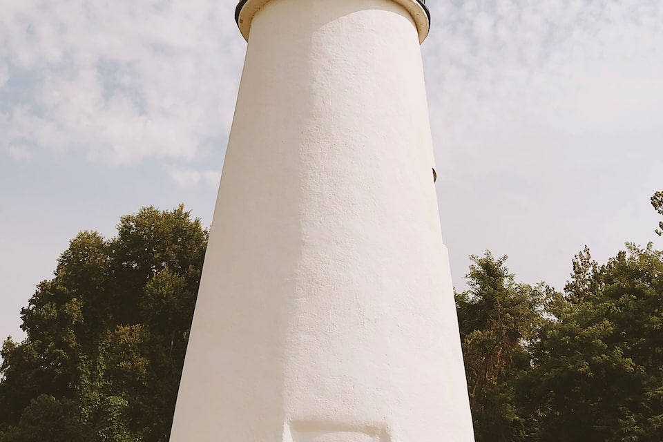 A white lighthouse with a black top and a small door at the base, surrounded by grass and trees.