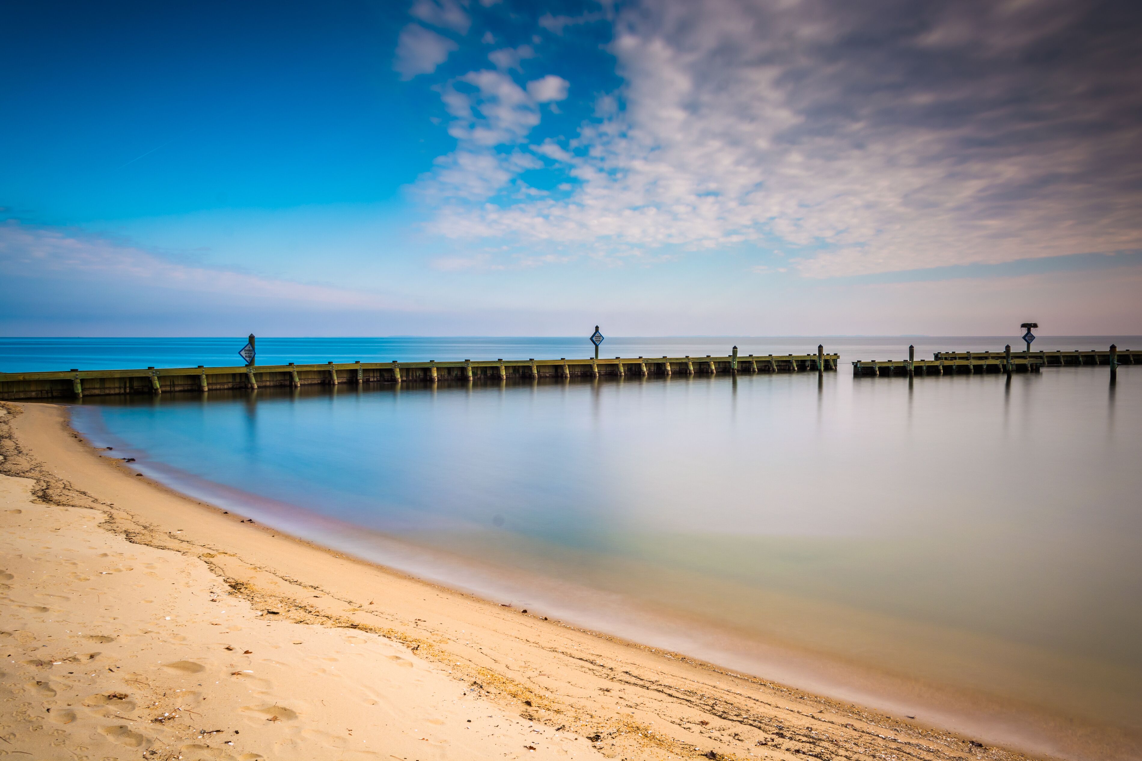A beach with a wooden pier extending into calm waters under a partly cloudy sky.