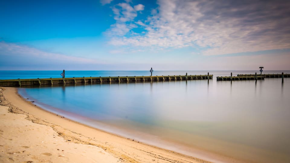 A beach with a wooden pier extending into calm waters under a partly cloudy sky.