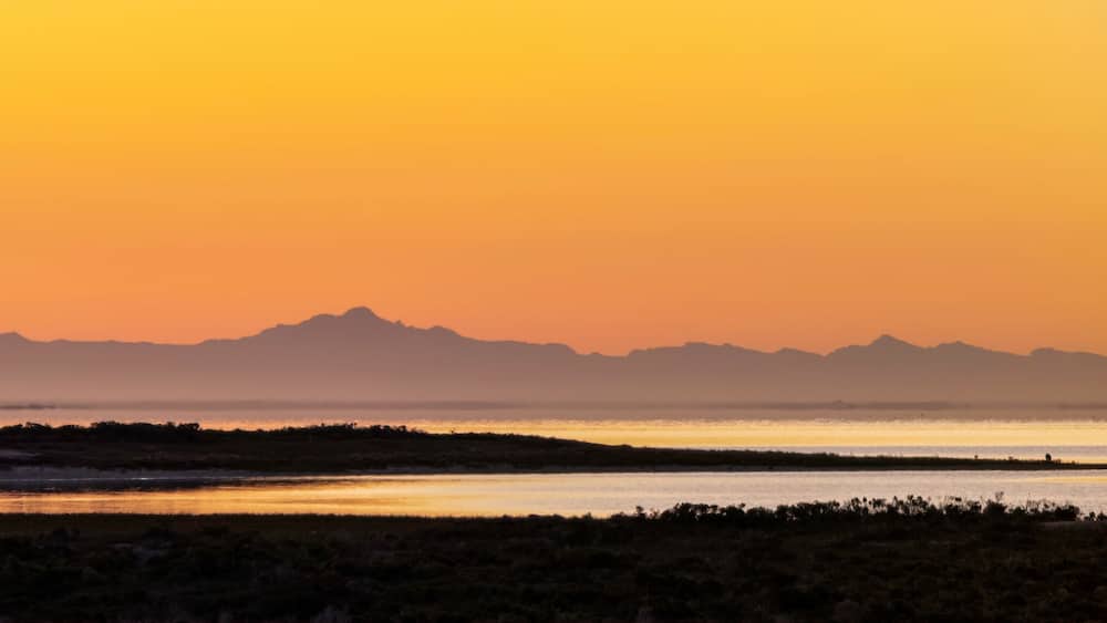 A sunset over a flat landscape with a water body and distant mountains.