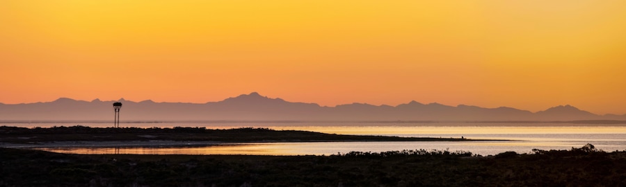 A sunset over a flat landscape with a water body and distant mountains.