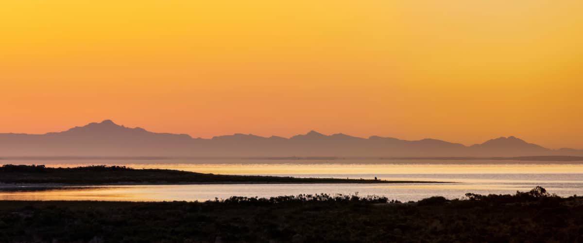 A sunset over a flat landscape with a water body and distant mountains.
