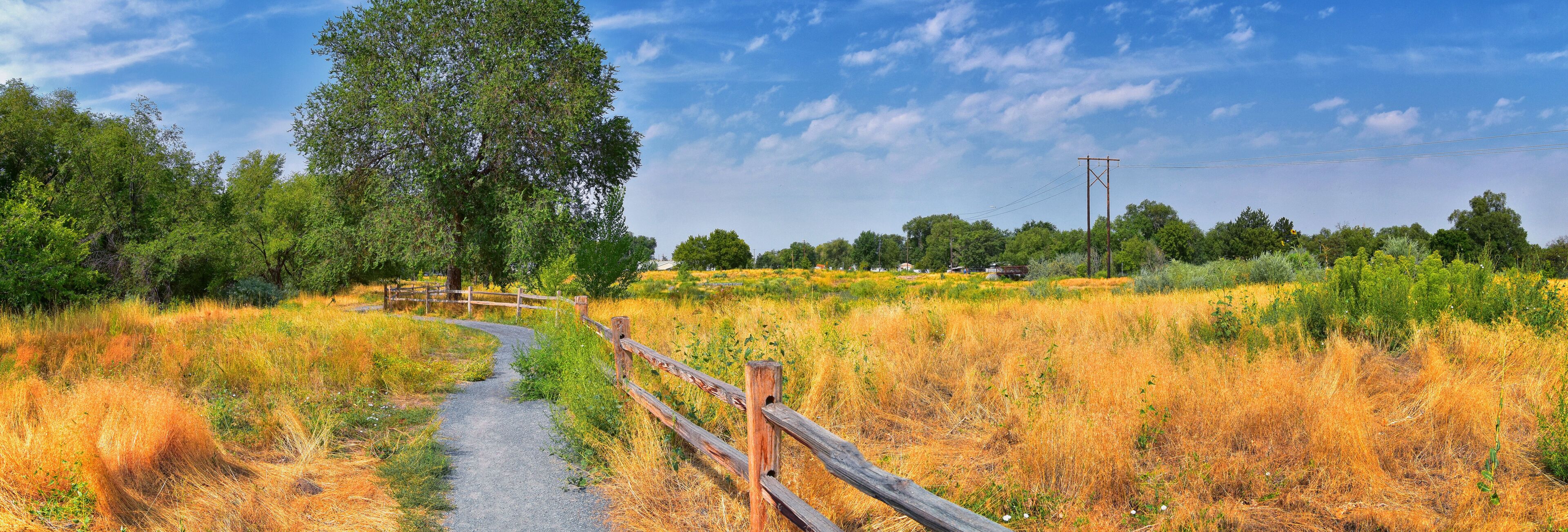 A winding gravel path through a grassy field with a wooden fence, surrounded by trees and a clear blue sky.