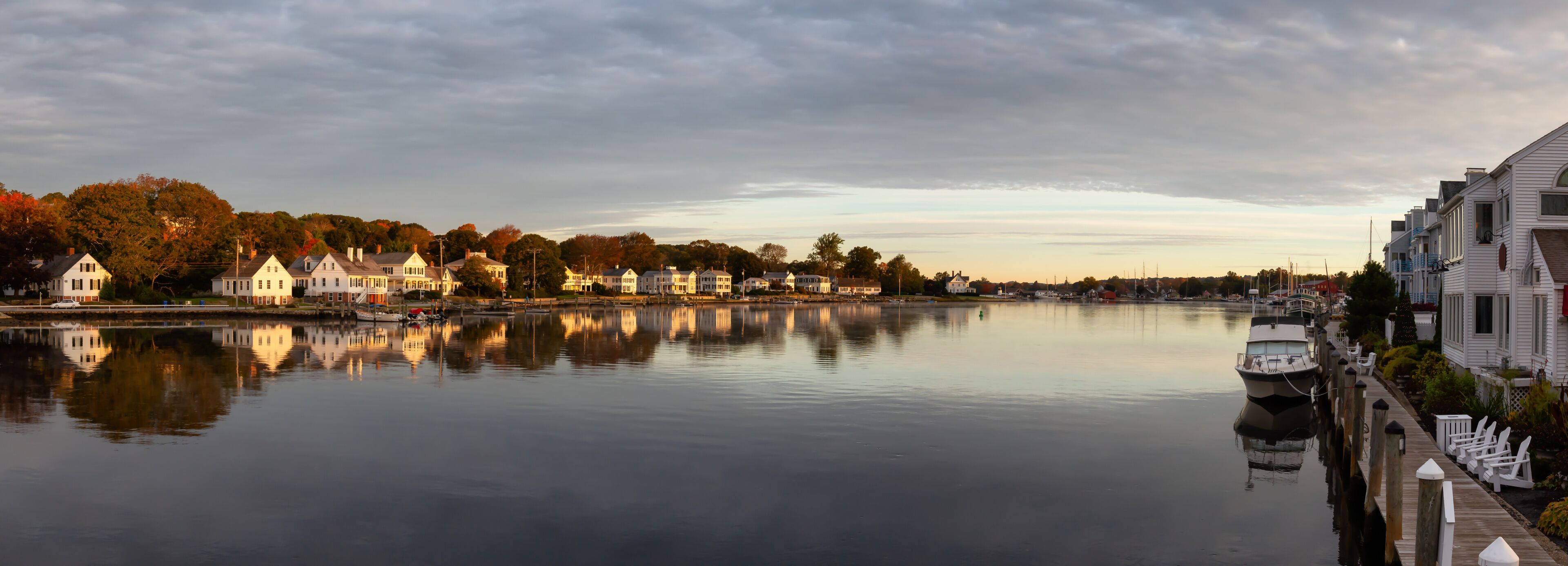 A calm river with houses reflecting on the water, surrounded by trees with autumn foliage.