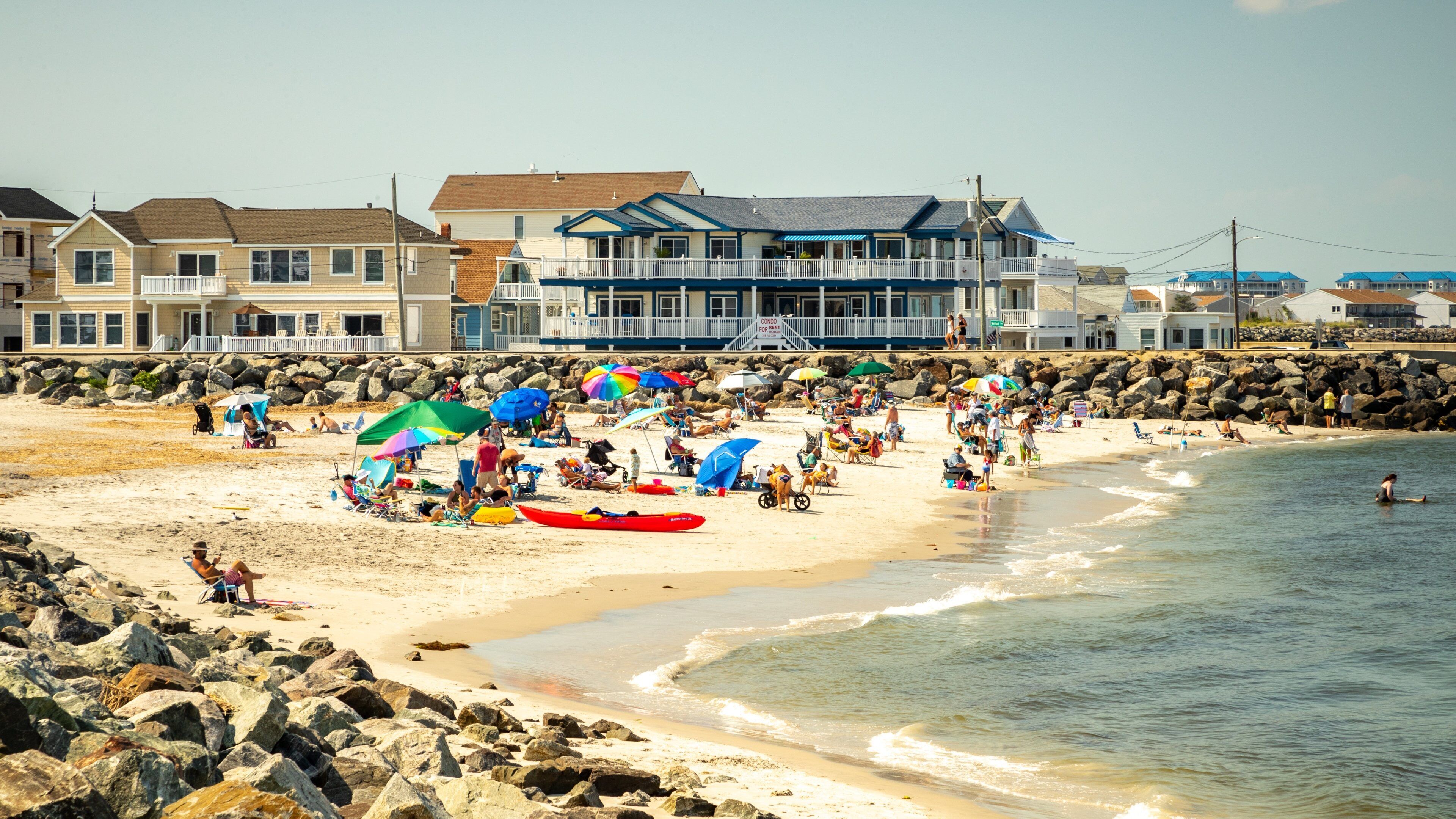 A crowded beach with people under colorful umbrellas, a red kayak, and houses in the background.