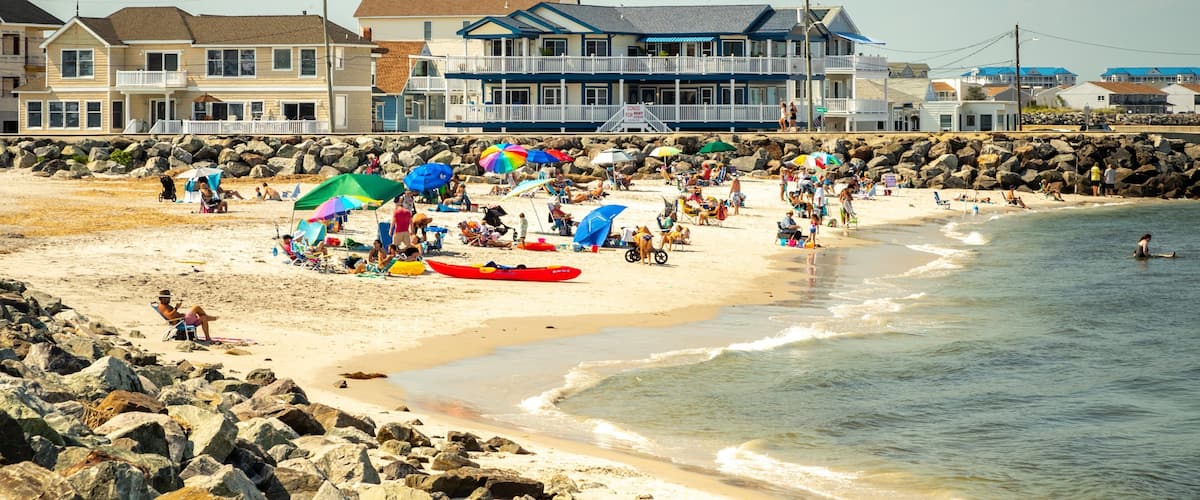 A crowded beach with people under colorful umbrellas, a red kayak, and houses in the background.