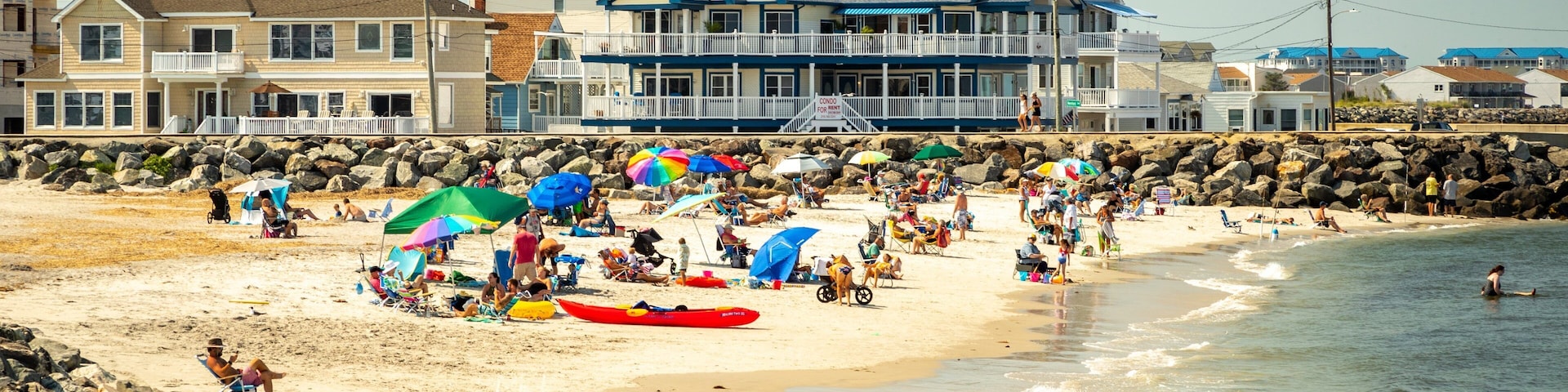 A crowded beach with people under colorful umbrellas, a red kayak, and houses in the background.