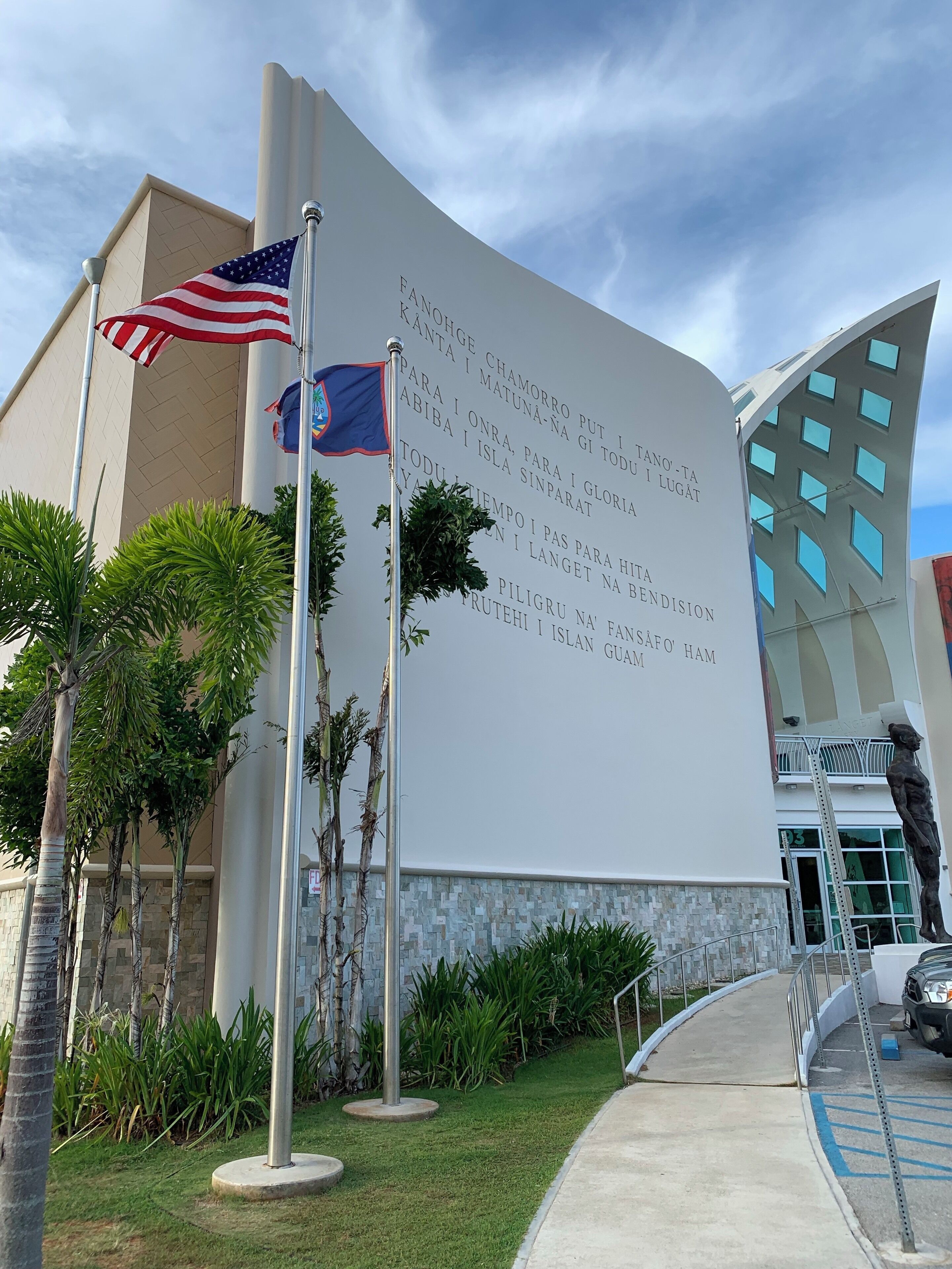 A building with a large white wall displaying text in multiple languages, two flags (American and another flag), and a statue of a woman.