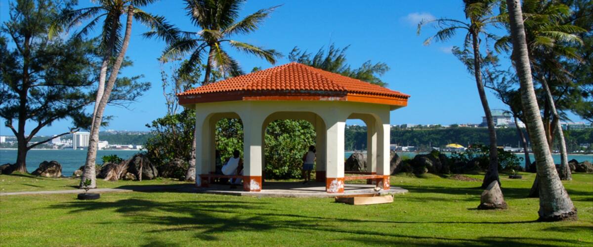 A gazebo with a red roof and white walls, surrounded by grass and palm trees.
