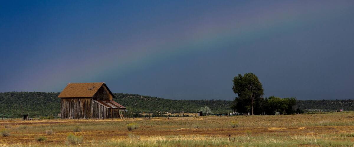 A rustic barn in a field with a rainbow in the sky.