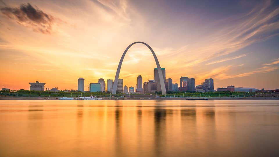 A city skyline with a prominent arch bridge reflecting in the water during sunset.