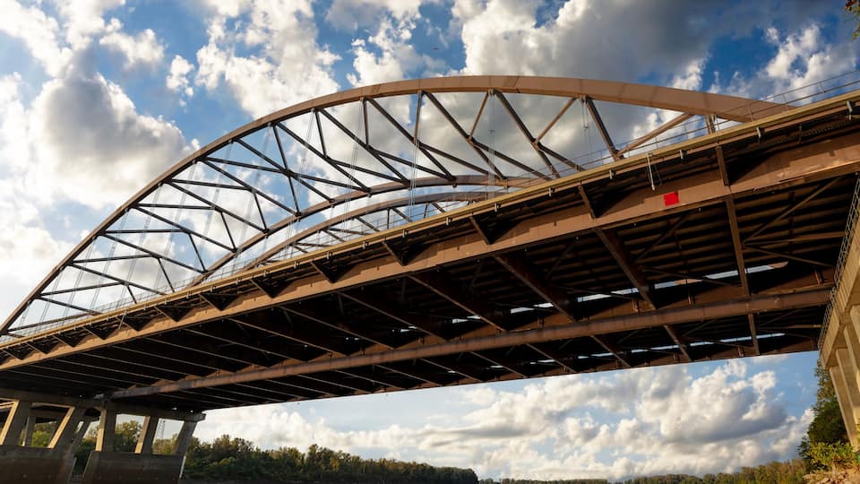A large steel bridge with a curved arch, surrounded by trees and a partly cloudy sky.
