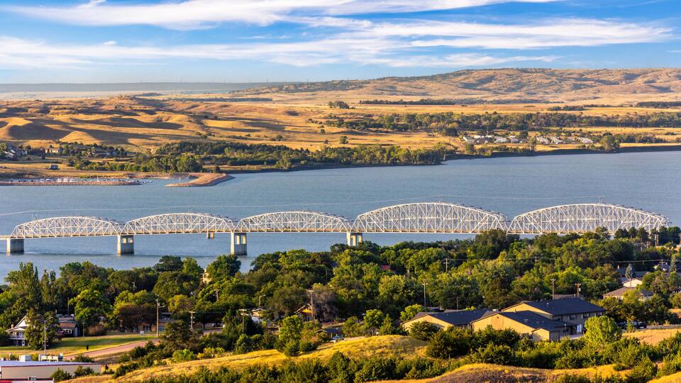 A large river with a bridge spanning across it, surrounded by hills and a small town with houses.