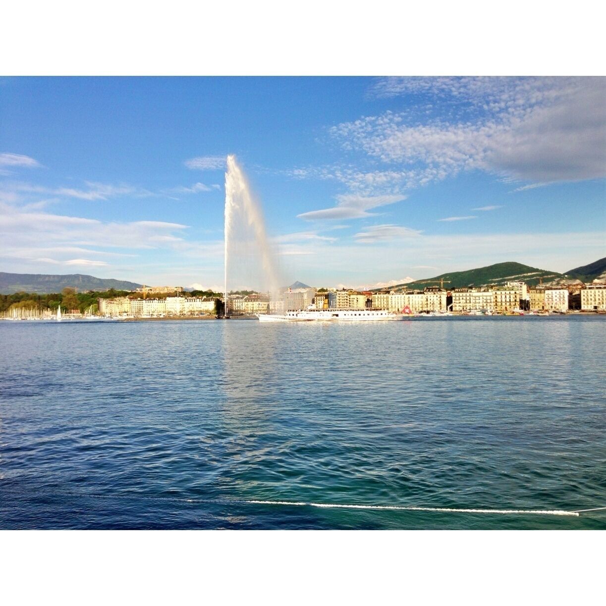 Une vue urbaine sur le front de mer, avec une fontaine imposante, des bâtiments le long de la côte et un ciel bleu dégagé.