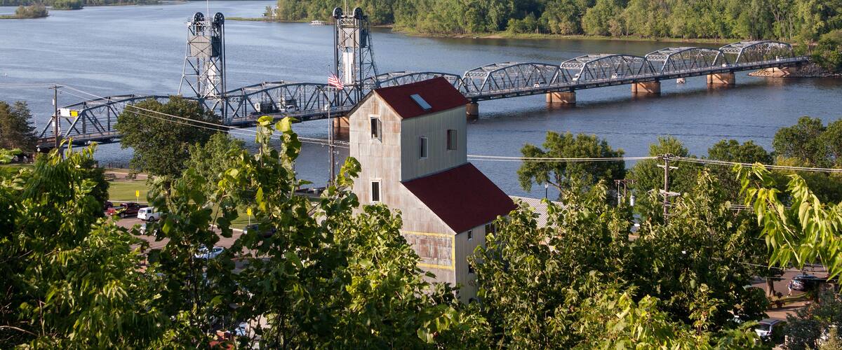 A steel bridge over a river, surrounded by lush greenery and a small building with a red roof.