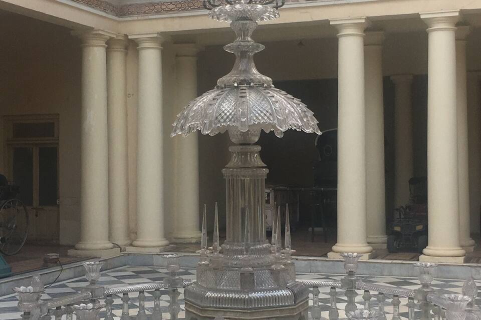 A grand fountain with intricate designs, surrounded by a checkered floor and columns in a spacious courtyard.