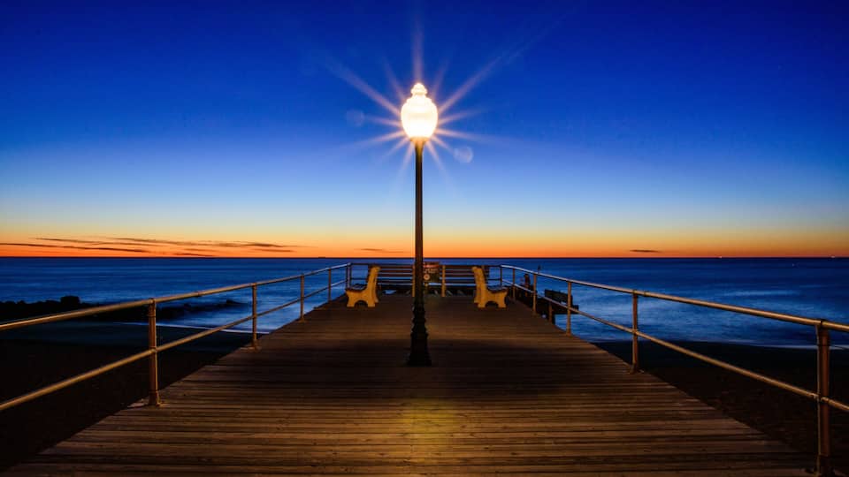 A wooden pier extending into the ocean at sunset with a street lamp at the end.
