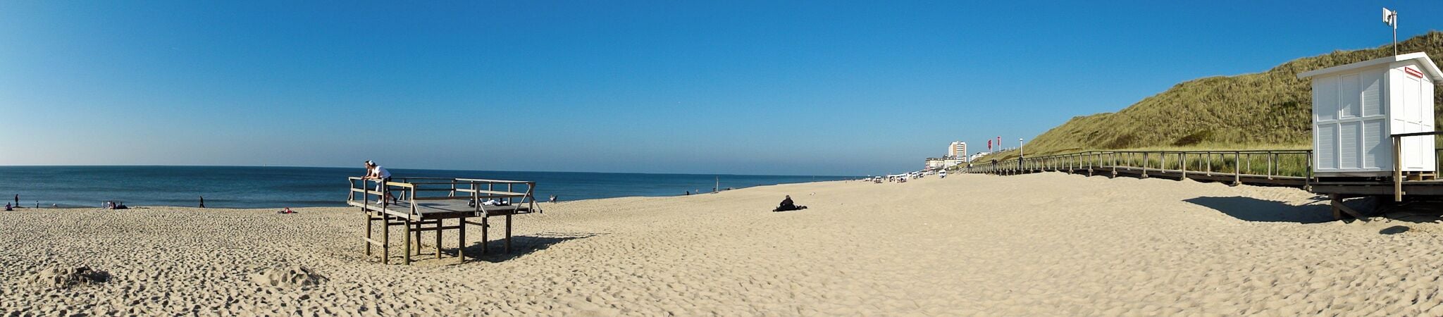 A sandy beach with a wooden bench, a lifeguard tower, and a grassy hill in the background.