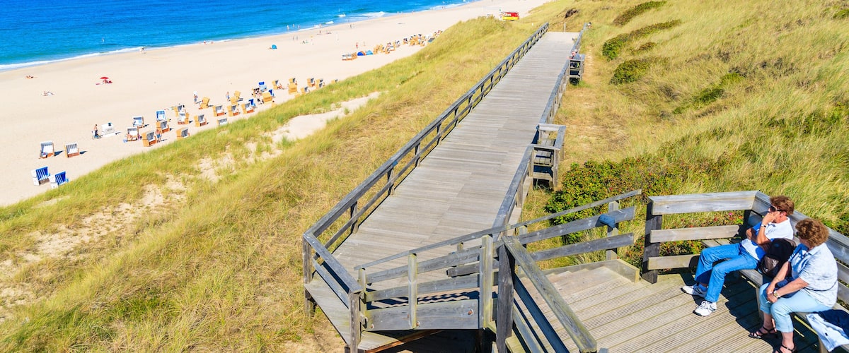 A beach with a wooden boardwalk, beach umbrellas, and people sitting on a bench.