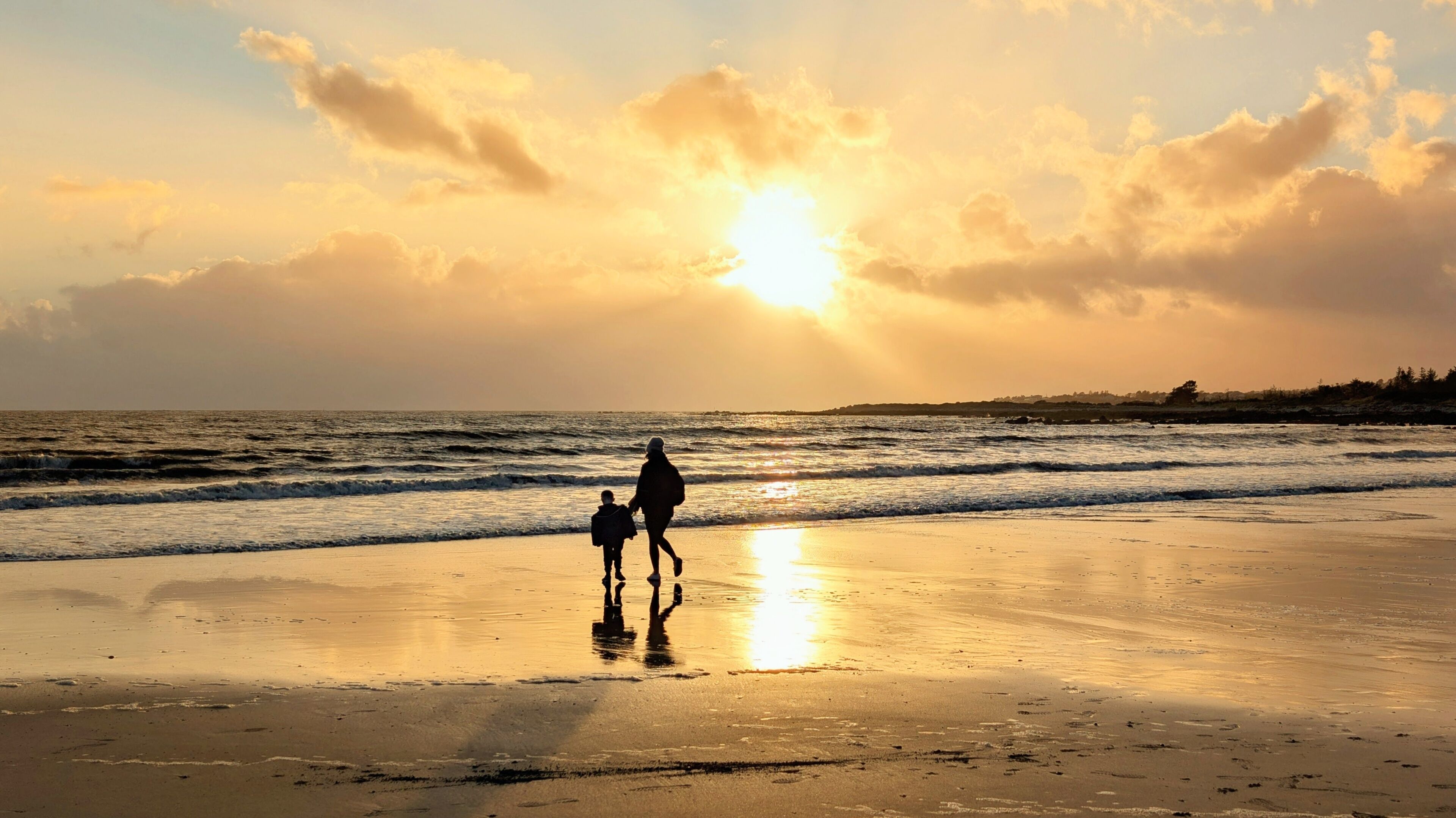 A beach with a sunset, waves, and two people walking.