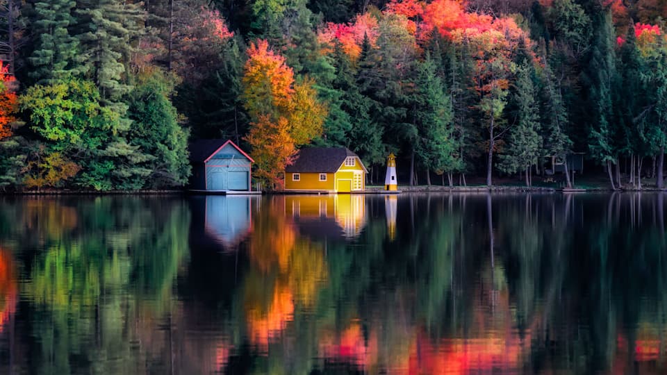 Autumn colors reflect in the waters at Old Forge Pond in NY. A Boat House, shed and lighthouse on the opposite shore look beautiful in Autumn Colors.