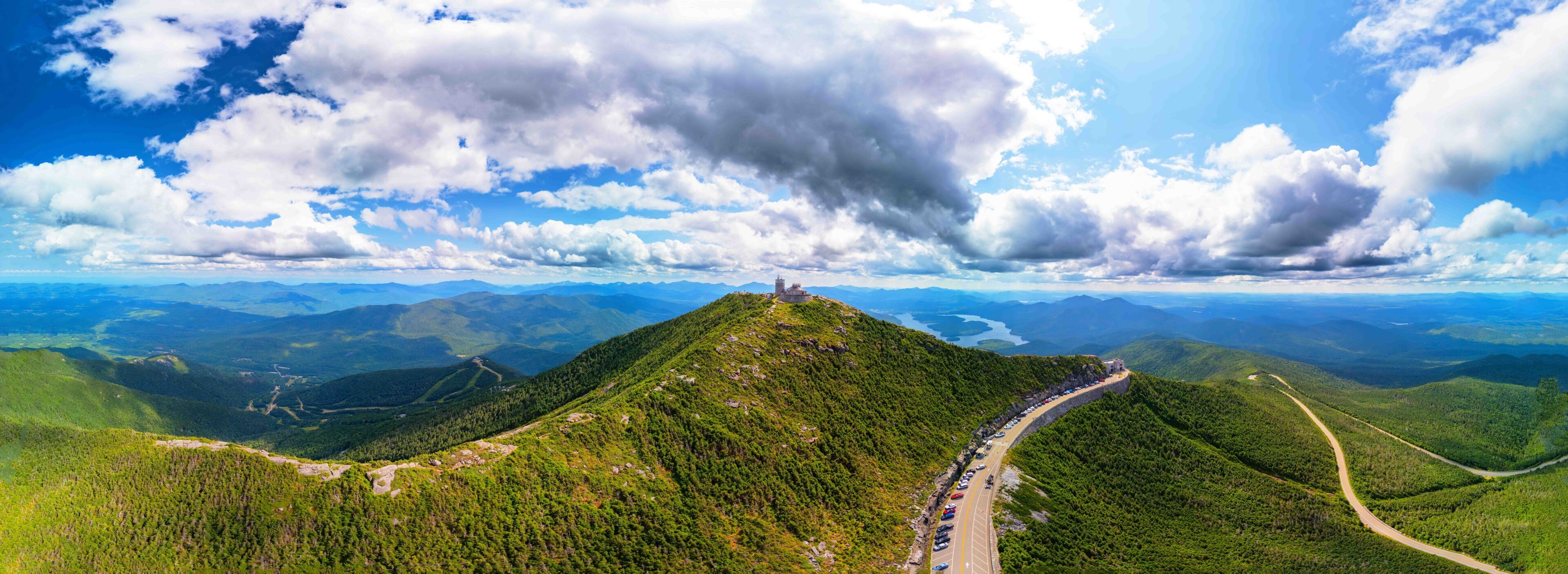 A mountain with a road leading to a castle-like structure on top, surrounded by lush green forests and distant mountains.