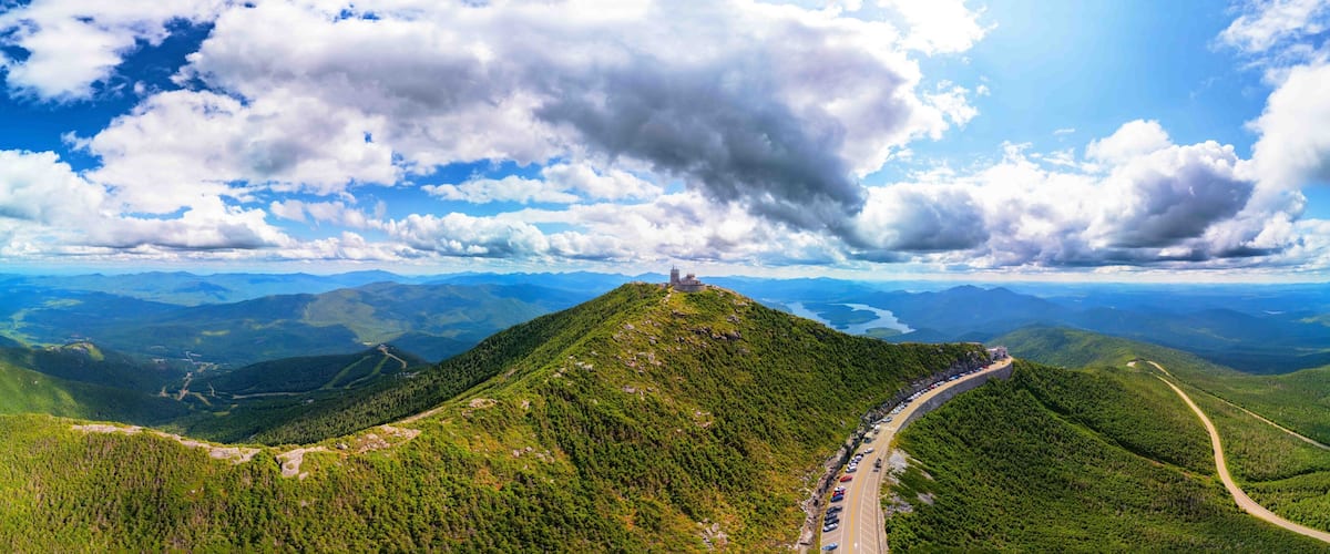 A mountain with a road leading to a castle-like structure on top, surrounded by lush green forests and distant mountains.