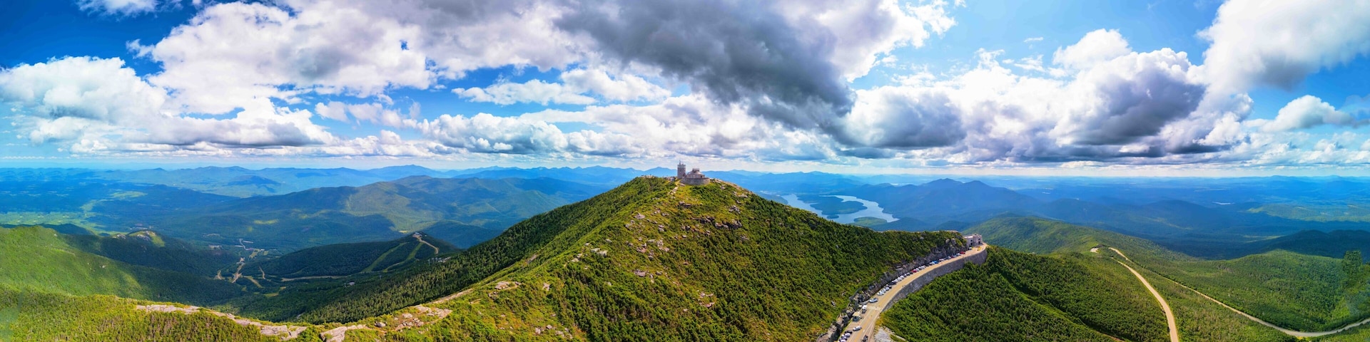 A mountain with a road leading to a castle-like structure on top, surrounded by lush green forests and distant mountains.