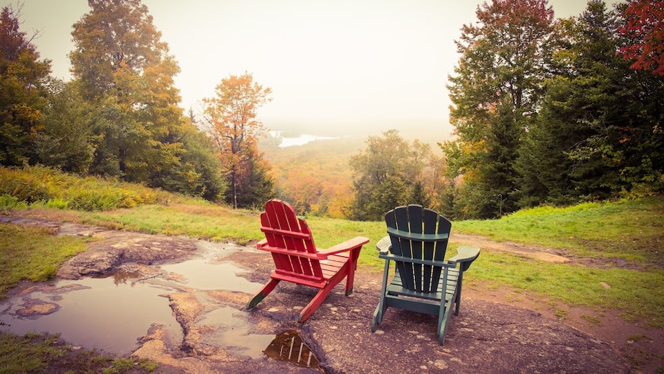 Two Adirondack chairs on a rocky surface with a view of a forest and a body of water in the distance.