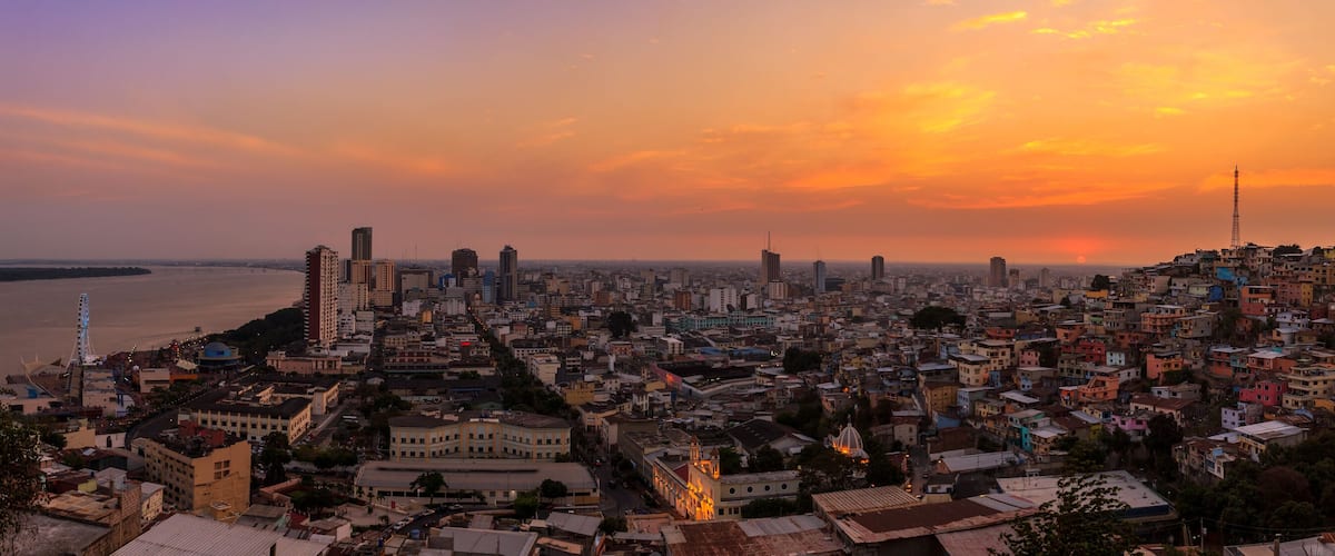 A cityscape at sunset with a river in the foreground and a mix of high-rise and low-rise buildings.