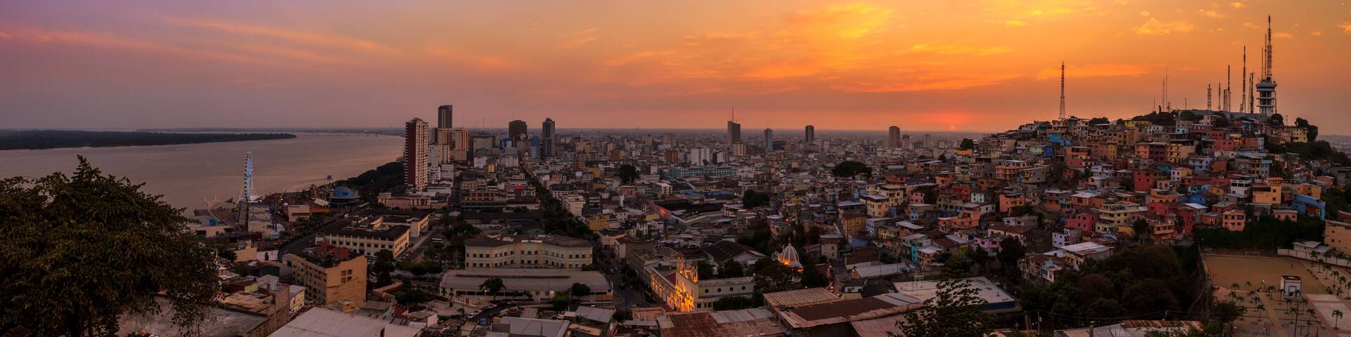 A cityscape at sunset with a river in the foreground and a mix of high-rise and low-rise buildings.